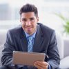 Businessman sitting on sofa using his tablet smiling at camera in the office